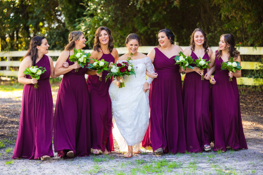 Bride and bridesmaids in burgundy dresses laugh during formal posed photos at a fall wedding in Charleston.