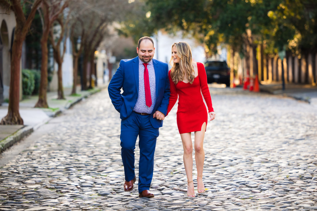 A couple walks down a cobblestone street holding hands and smiling during their engagement session in downtown Charleston, South Carolina.