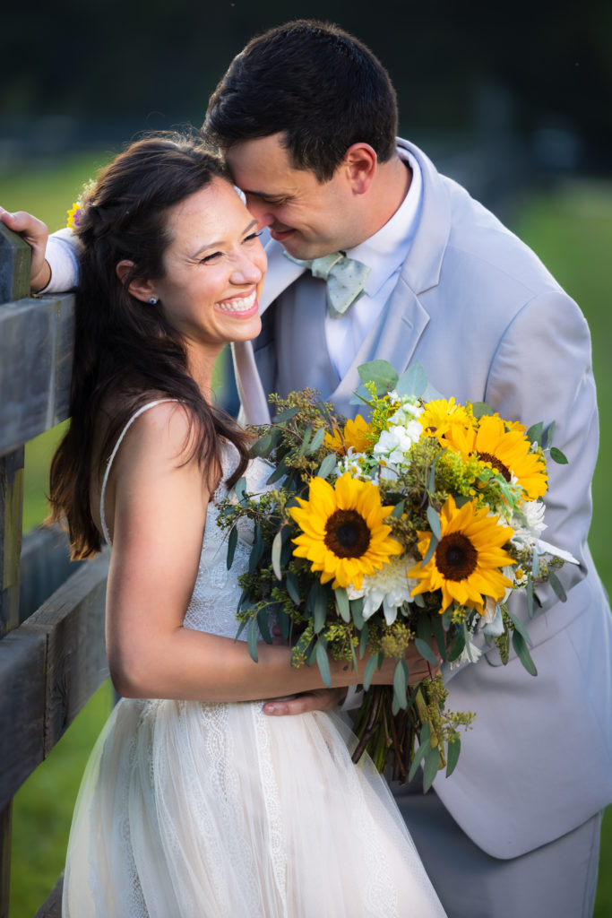 A couple leans against a fence with bride holding a sunflower bouquet at a fall wedding Charleston.